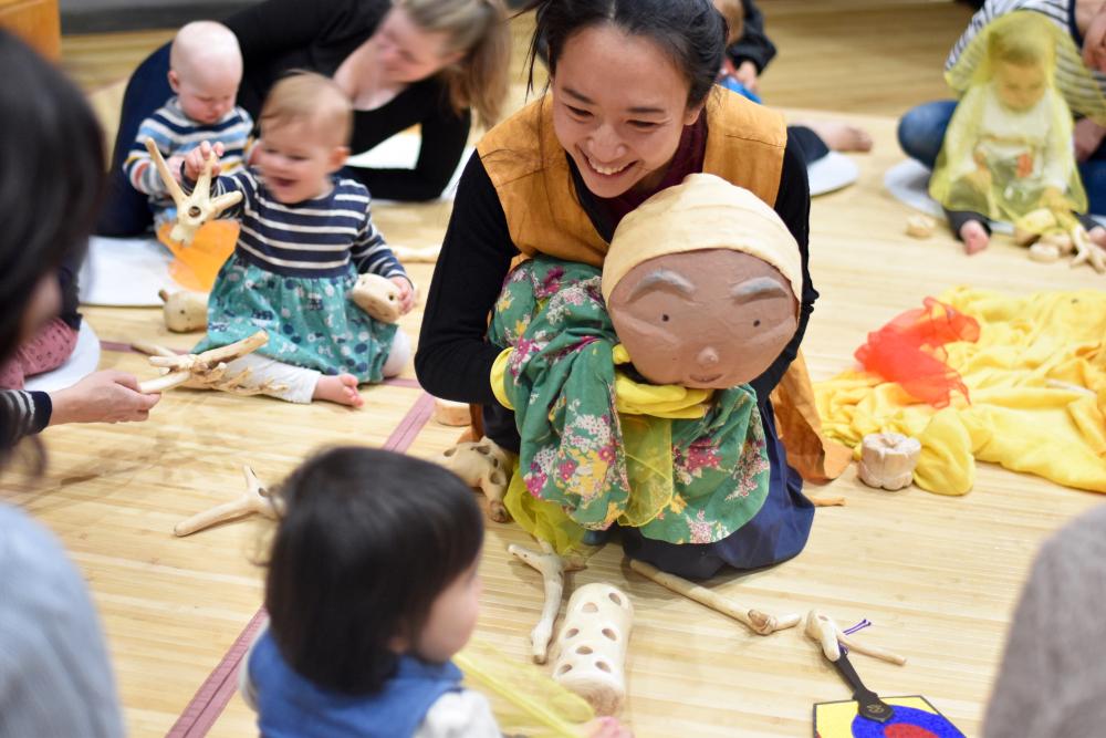 An artist sitting on the floor holding up a puppet in front of a child