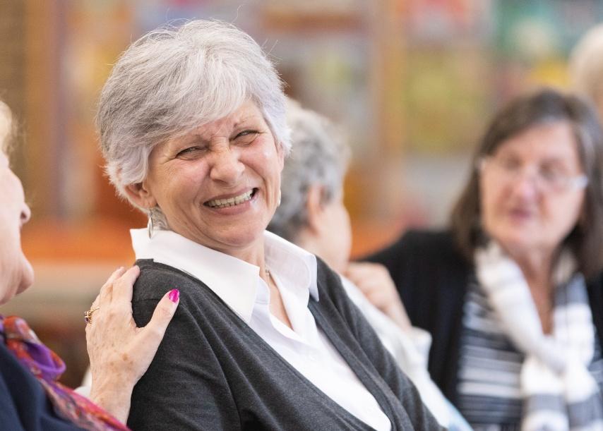 An older woman smiles at a woman sitting beside her, who has placed a friendly hand on her shoulder. The other woman's face is not visible in the photo.