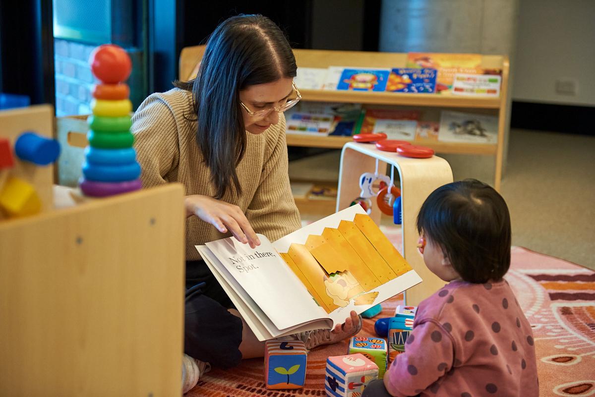A female-presenting person reads a picture book to a young toddler. They are sitting on the floor and are surrounded by books and toys.