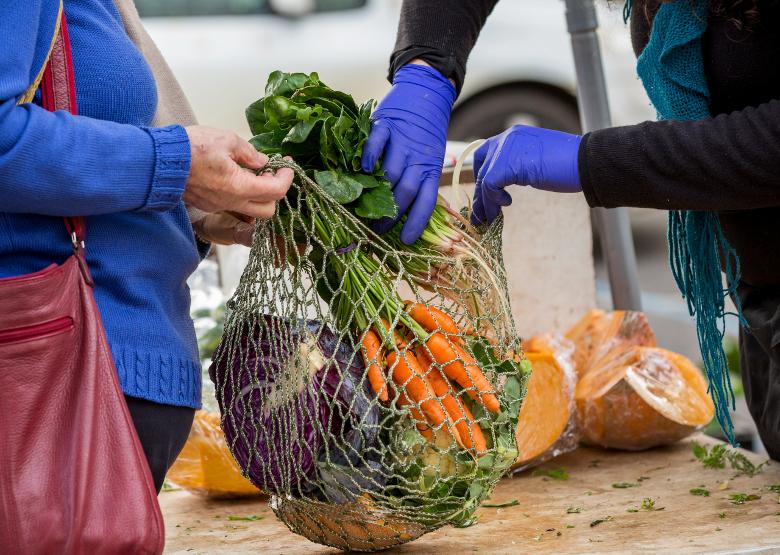 A person in a blue sweater is holding a netted bag full of vegetables, which is being filled with produce by a vendor. Only the two people's hands are visible. The vendor is wearing blue gloves.