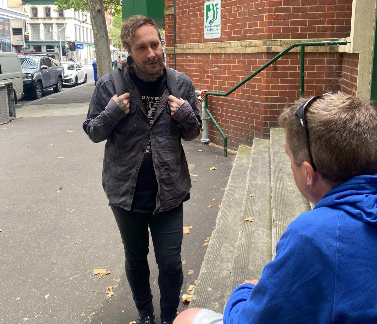 A man with neck tattoos, large earrings, wearing a jacket and backpack approaches another man sitting on a flight of outdoor steps. He is wearing a blue hoodie and sunglasses on his head.
