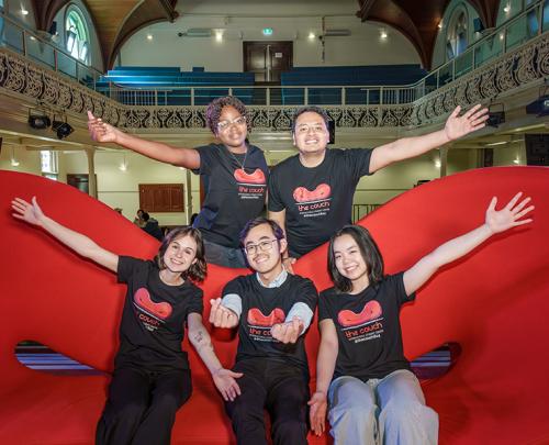 A group of five students wearing matching black “The Couch” T‑shirts pose for a photo on a large red couch, smiling and extending their arms enthusiastically.