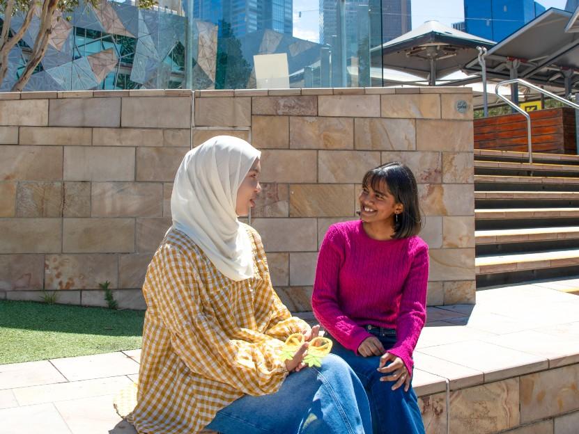 Two young women sit on a ledge in Federation Square, having a conversation with one another. The woman on the left is wearing a white hijab.