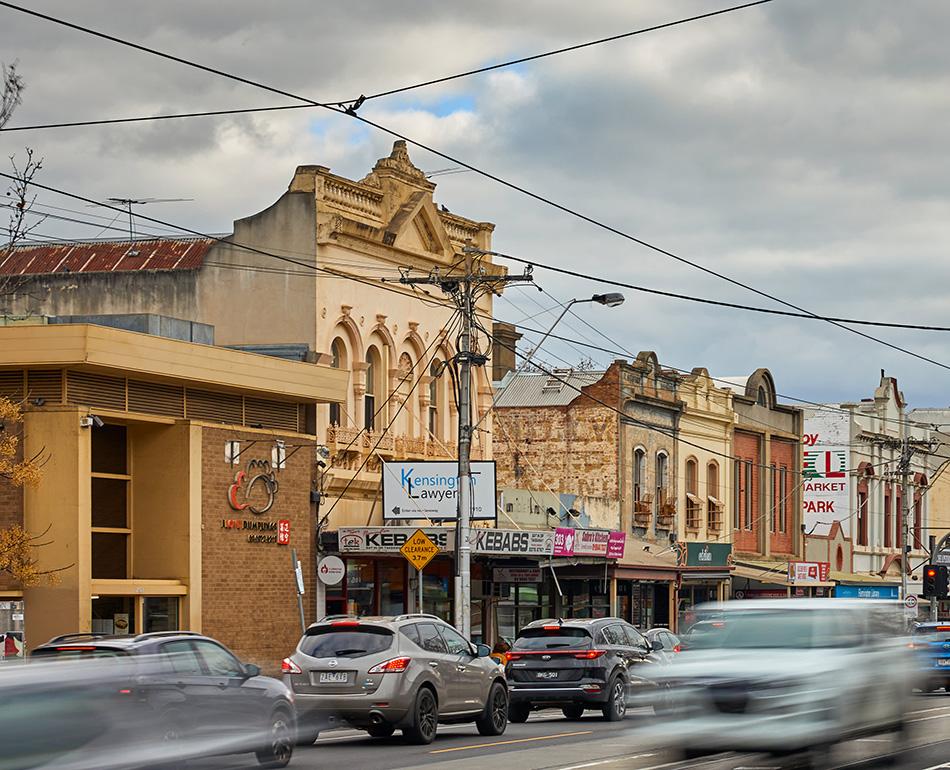 Shops and businesses in low-rise Victorian buildings on a busy street with cars and trams,