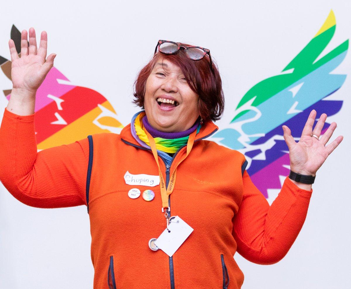 An older Asian woman with short red hair and glasses on her head smiles at the camera, holding her hands up. She is standing in front of rainbow wings which have been painted on the wall behind her. She is wearing an orange top and vest, an orange lanyard, two small badges on her chest and a nametag which reads " Chuping".