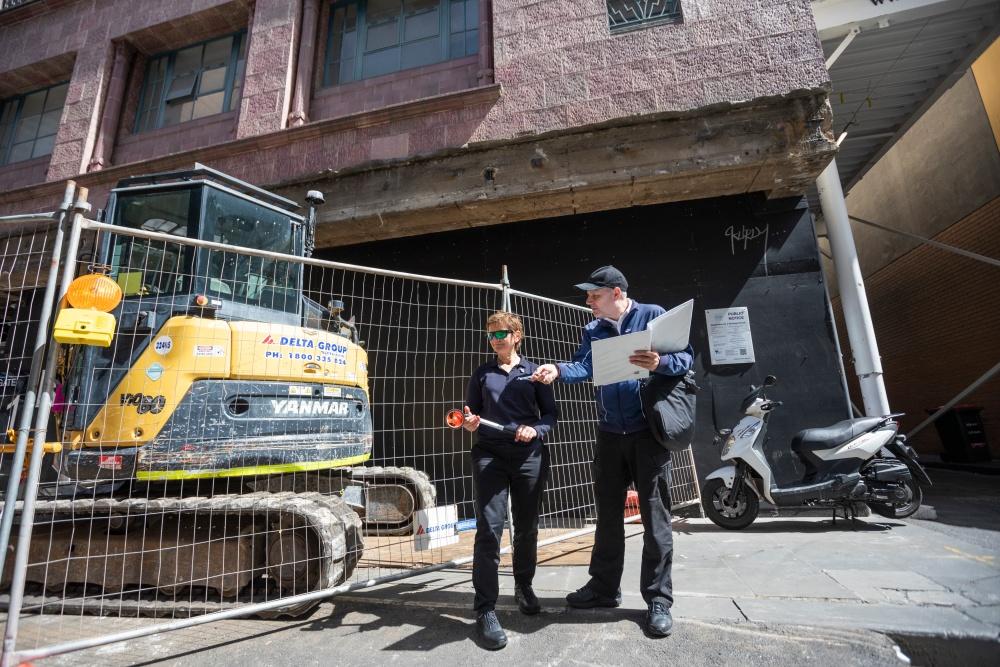 Two people with clipboards and a bulldozer behind temporary fencing parked on footpath