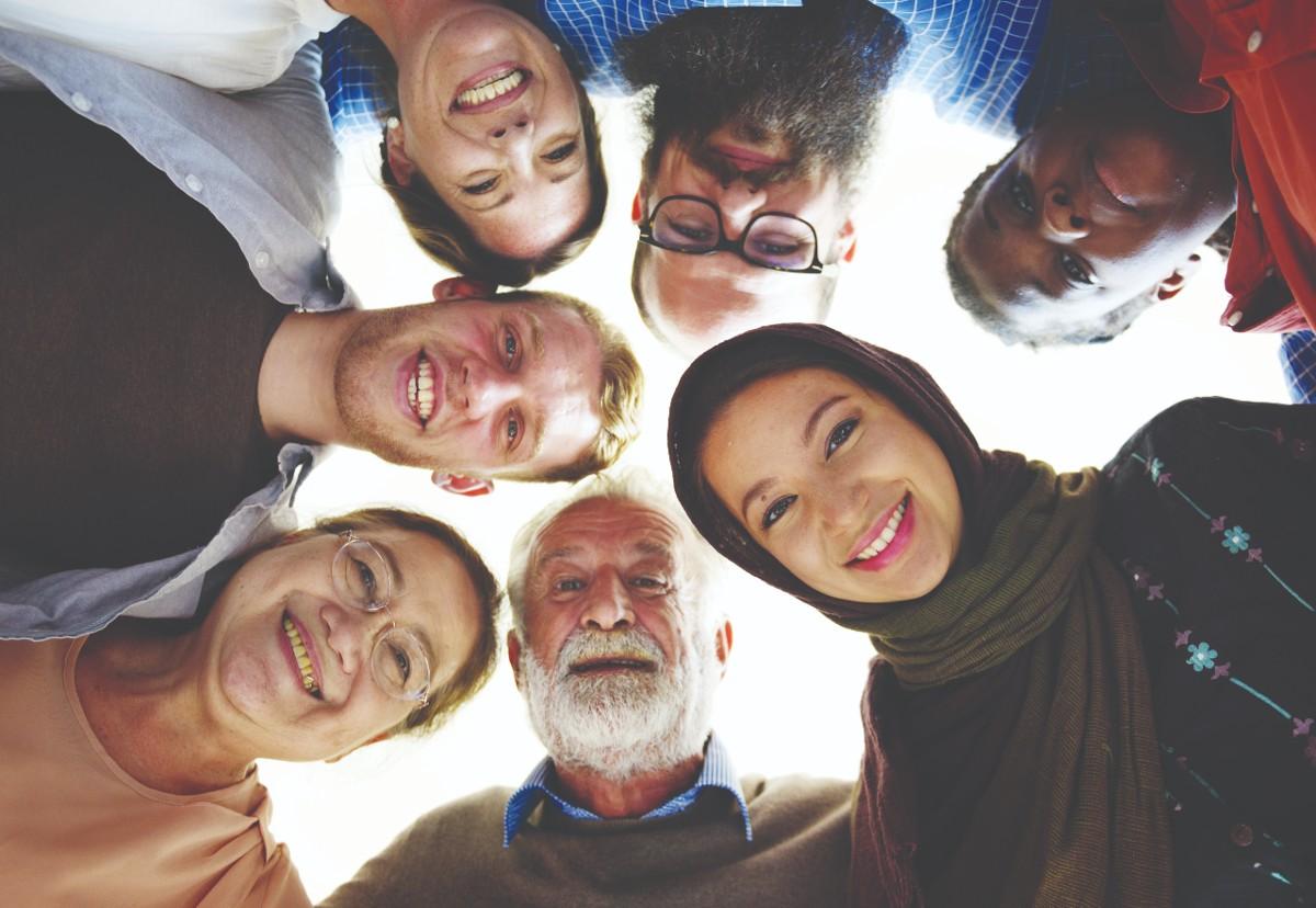 A group of seven people of different ages, genders and ethnicities stand with their heads close together, smiling down at the camera.