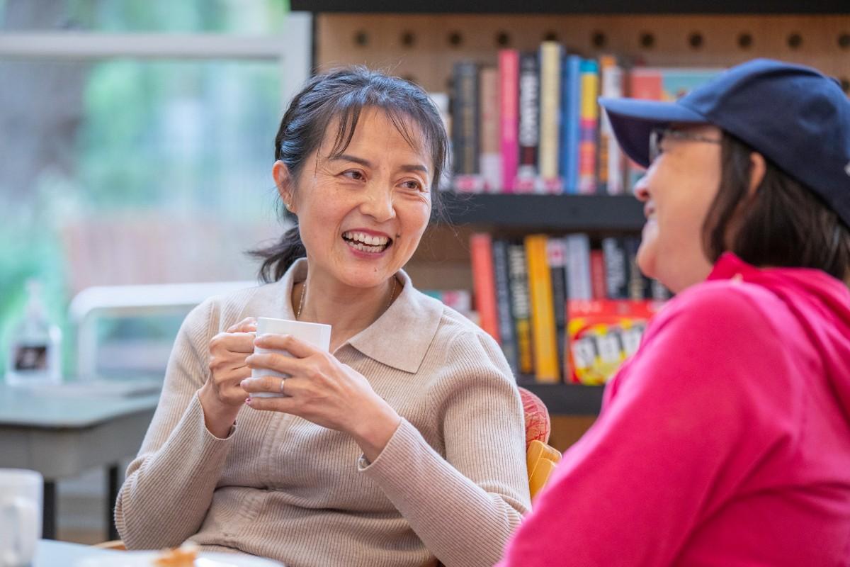 Two women sit with one another, laughing together. One woman holds a mug.
