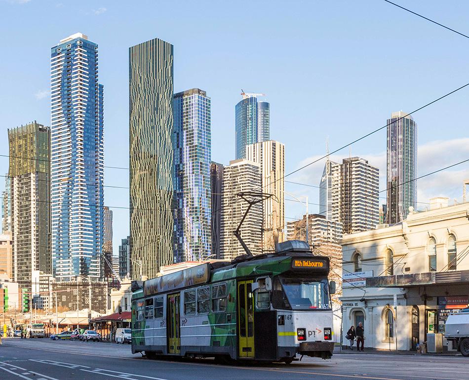 A tram travels on a North Melbourne street, with CBD high-rise buildings seen in the background.