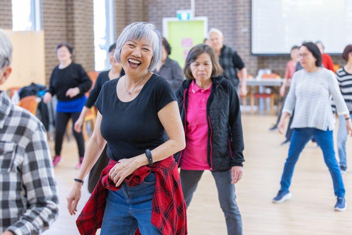 An older Asian woman with short grey hair smiles and dances with a group of other older Asian people in a large hall.