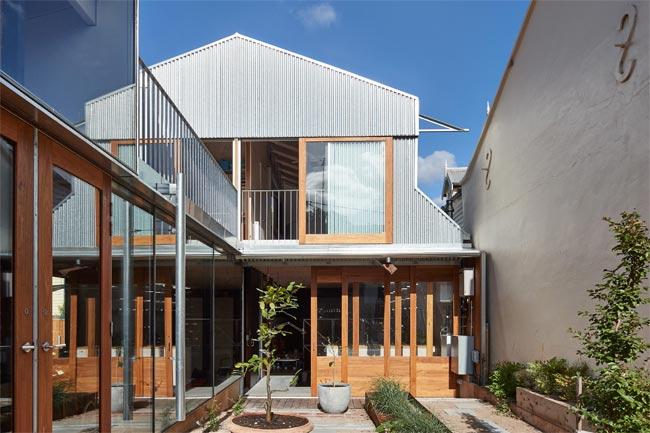 Back view of a modern two-storey house occupying space between heritage buildings. The house, overlooking a small courtyard, has corrugated metal cladding and large wood-framed windows and doors.