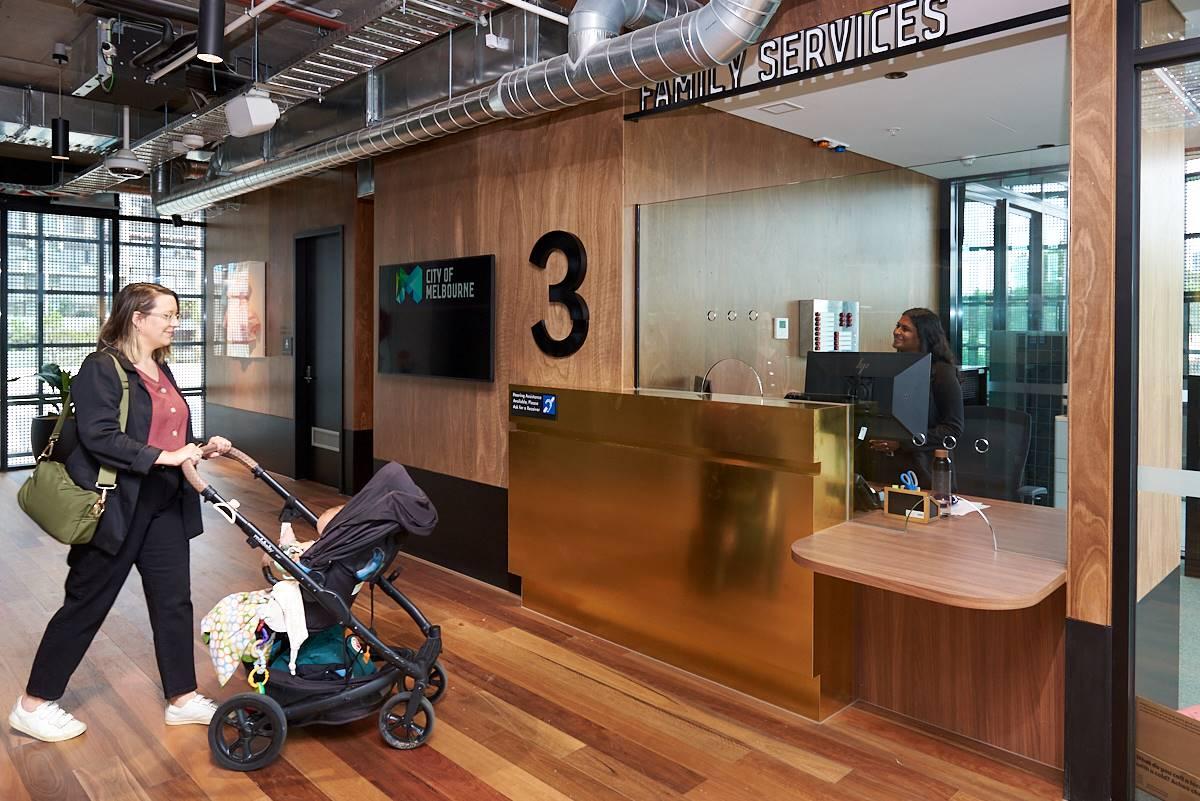 A mother pushes a pram with a baby towards a smiling receptionist, who stands at a desk underneath a sign that reads "Family Services"