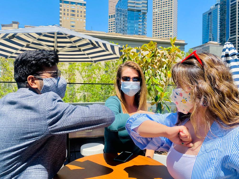 Three people are sat around a table at a rooftop bar, high-fiving each other with their elbows. They are all wearing masks.