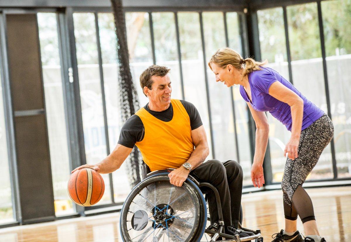 A man in a wheelchair is bouncing a ball in a game of basketball. A woman stands in front of him, trying to intercept the ball.