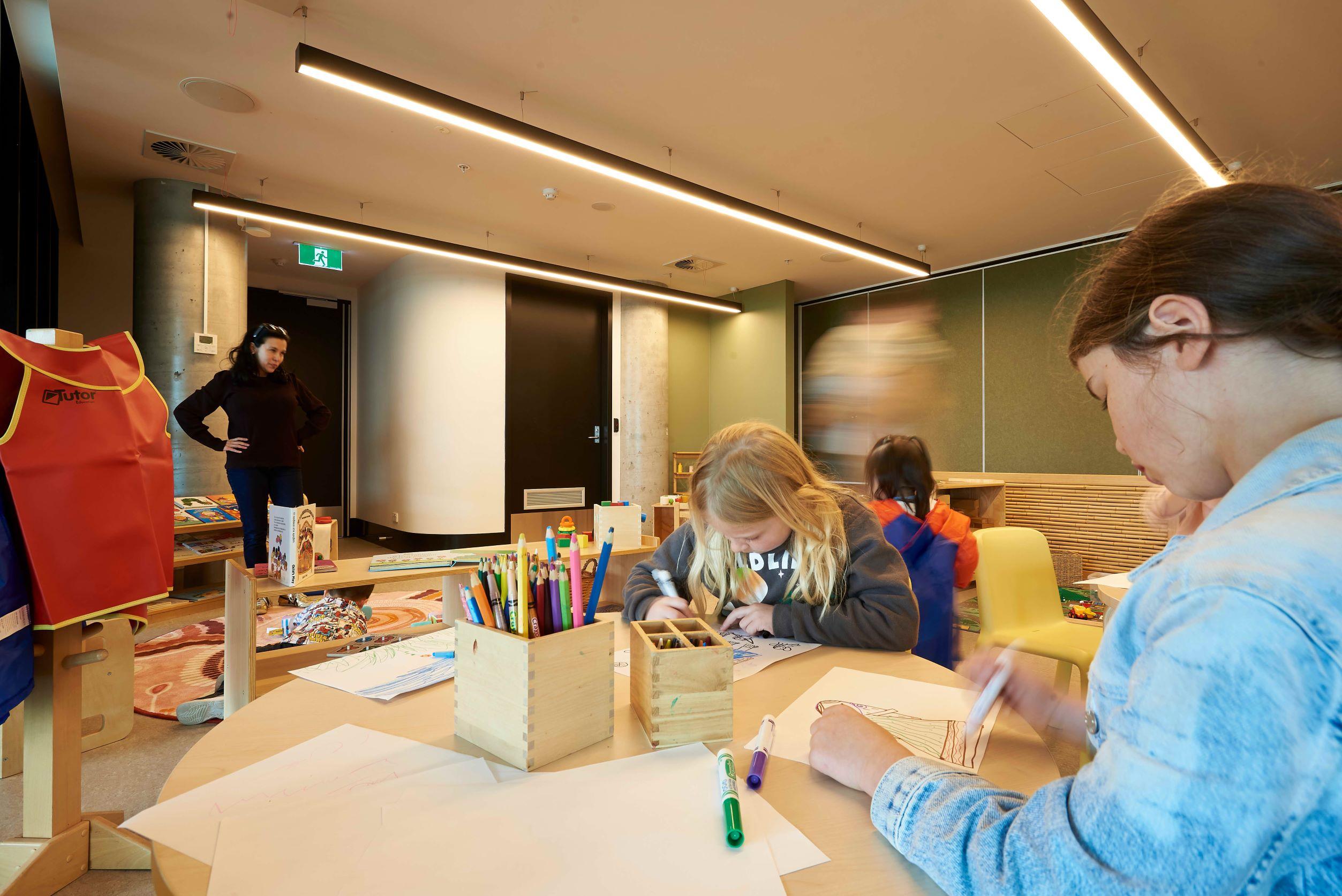 Three children sit around a table, drawing pictures using coloured textas. An adult supervises them.