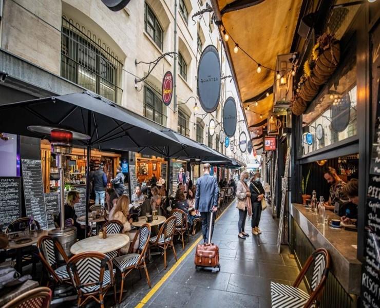 An inner-city Melbourne laneway, with eateries and tables and chairs running each side. Above each shop hangs a retro sign with their business name. A person wheeling a suitcase walks up the centre of the laneway, away from the viewer. 