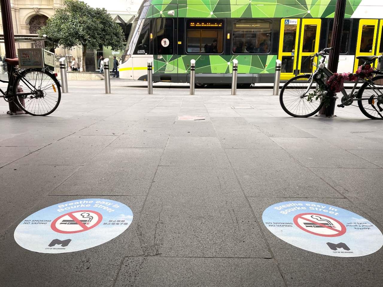 In the foreground of the image, two circular floor decals are depicted on the pavings along Bourke Street, Melbourne. They read: "Breathe Easy in Bourke Street" "No smoking No vaping". One has the messaging in English and Chinese (Simplified), the other has the message in English and Arabic. They both display the no smoking/no vaping symbol, the City of Melbourne logo atop a cloud background. In the background of the image, a tram passes by.