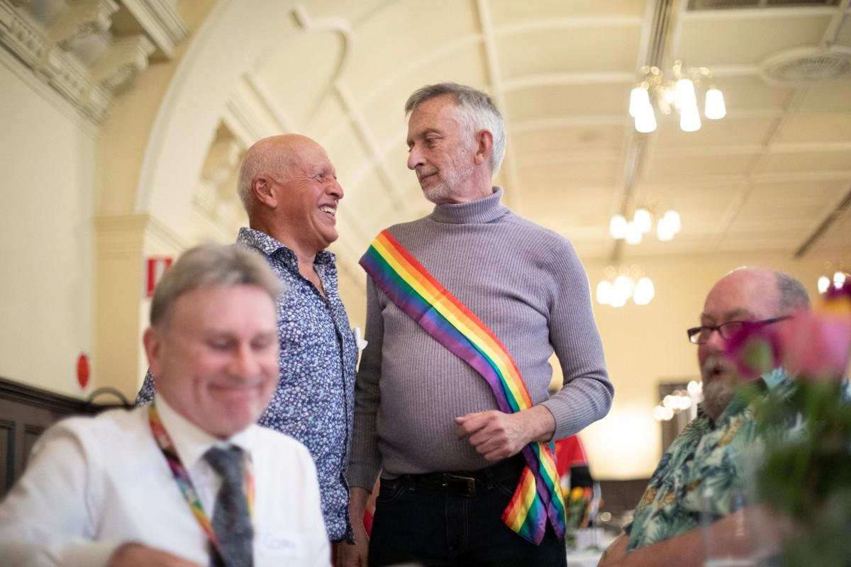 Two older men are standing in a room talking with one another. The man on the right is wearing a rainbow sash. In the foreground, two more men sit around a table.